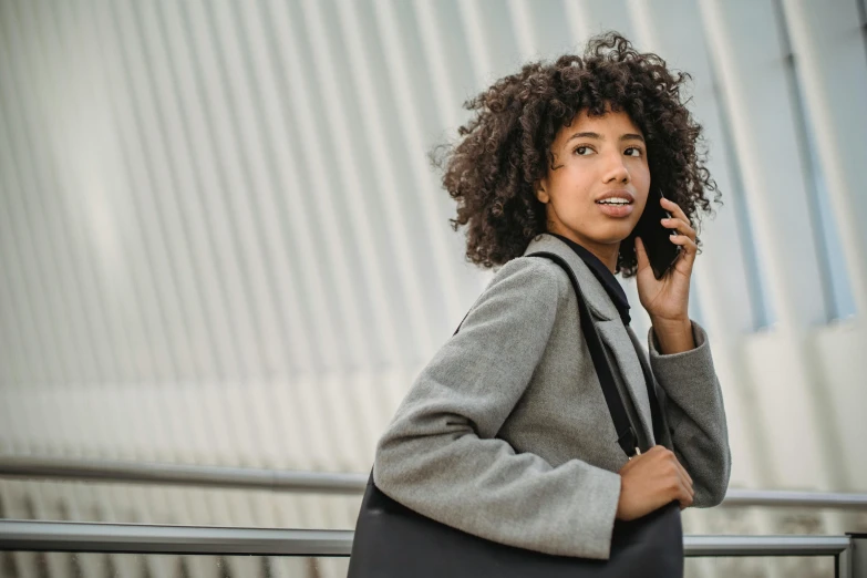 a woman with curly hair talking on a cell phone, pexels contest winner, holding a briefcase, grey turtleneck coat, black girl, with a backpack