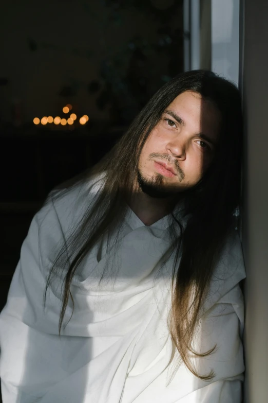 a man with long hair leaning against a wall, an album cover, inspired by Serhii Vasylkivsky, reddit, wearing white pajamas, headshot photo, standing in a dimly lit room, gif