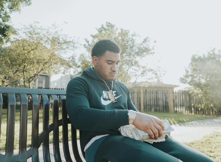 a man sitting on top of a wooden bench, rapper jewelry, jayson tatum, in a suburb, cash