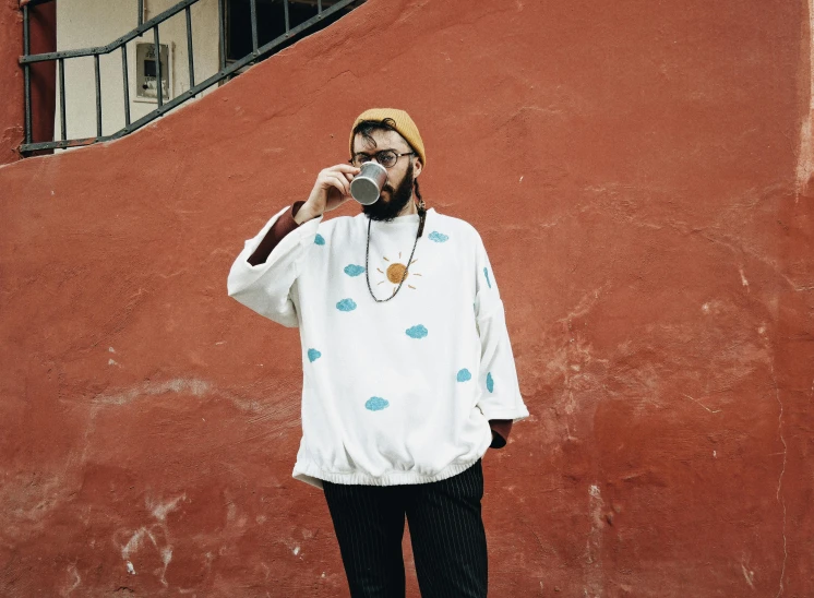 a man standing in front of a red wall talking on a cell phone, an album cover, pexels contest winner, graffiti, drinking a cup of coffee, uniform off - white sky, poncho, hipster