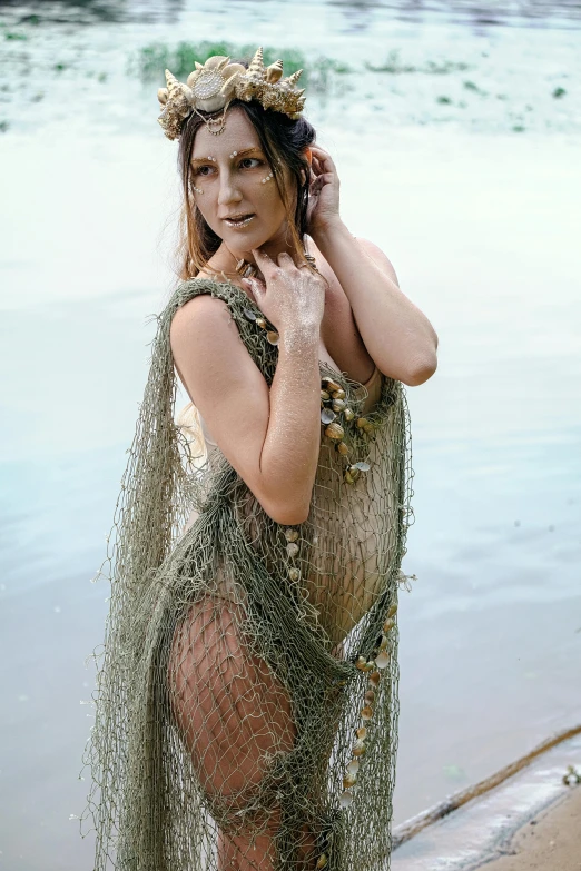 a woman standing on a beach next to a body of water, fishnet clothes, body draped in moss, wearing seashell attire, promo shoot