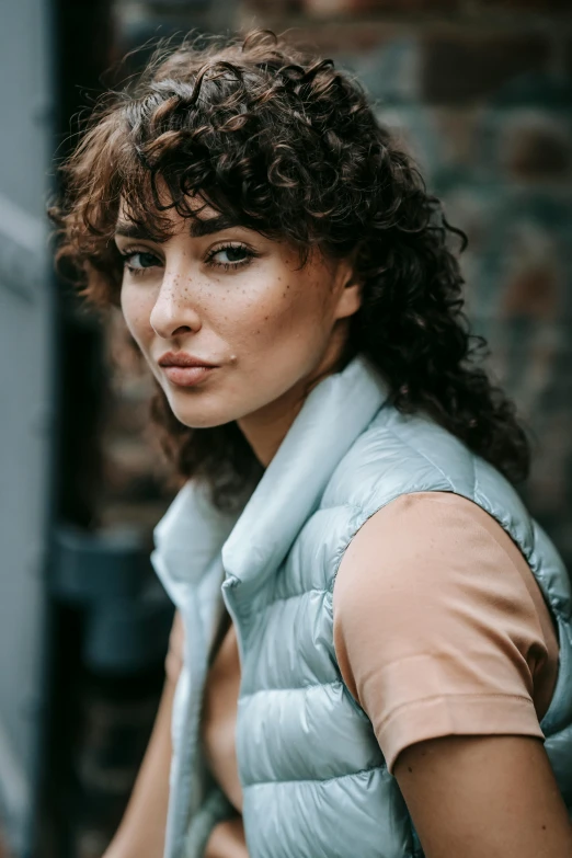 a woman with curly hair wearing a blue vest, by Nina Hamnett, trending on pexels, middle eastern skin, grey jacket, caramel. rugged, influencer