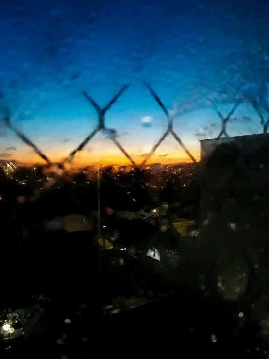 a view of a city through a chain link fence, an album cover, inspired by Elsa Bleda, ((sunset)), window ( rain ), low quality photo, gopro photo