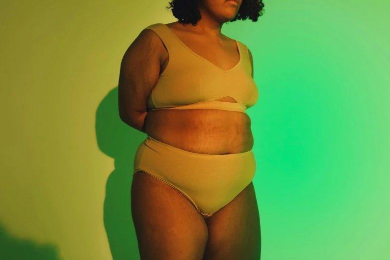 a woman in a tan bikini standing in front of a green wall, trending on pexels, morbidly obese, ( ( brown skin ) ), soft luminescent glow, nettie wakefield