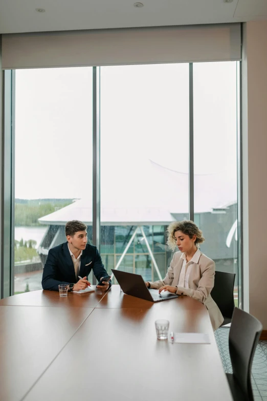 a couple of people sitting at a table with a laptop, in a meeting room, large windows, woman in business suit, lake view