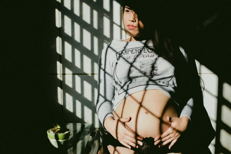 a pregnant woman standing in front of a window, a photo, pexels contest winner, sitting in a prison, belly button showing, half asian, female image in shadow