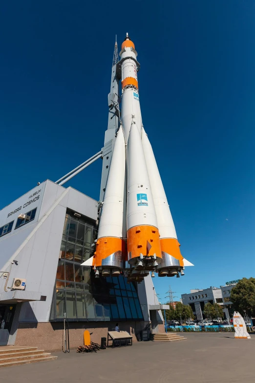 an orange and white rocket on display in front of a building, blue sky, vessels, over the shoulder, manuka