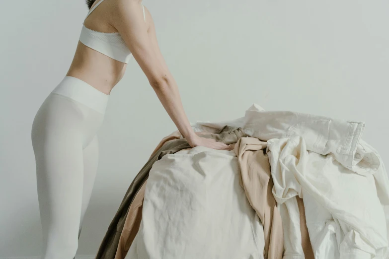 a woman standing next to a pile of clothes, by Emma Andijewska, pexels contest winner, figuration libre, white bed, taupe, background image, bending over