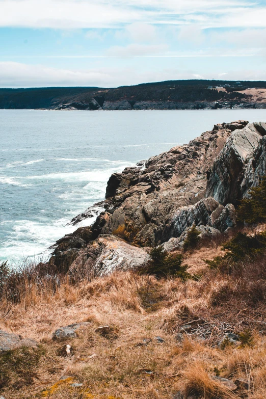 a man standing on top of a cliff next to the ocean, by Bascove, trending on pexels, les nabis, rocky grass field, maple syrup sea, slate, top of the hill