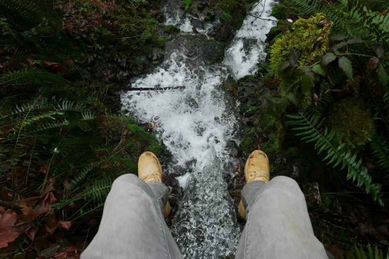 a person standing in front of a waterfall, legs taking your pov, sitting on the ground, as seen from the canopy, wearing boots