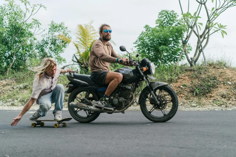 a man riding a motorcycle next to a woman on a skateboard, islandpunk, profile image, bali, outside on the ground