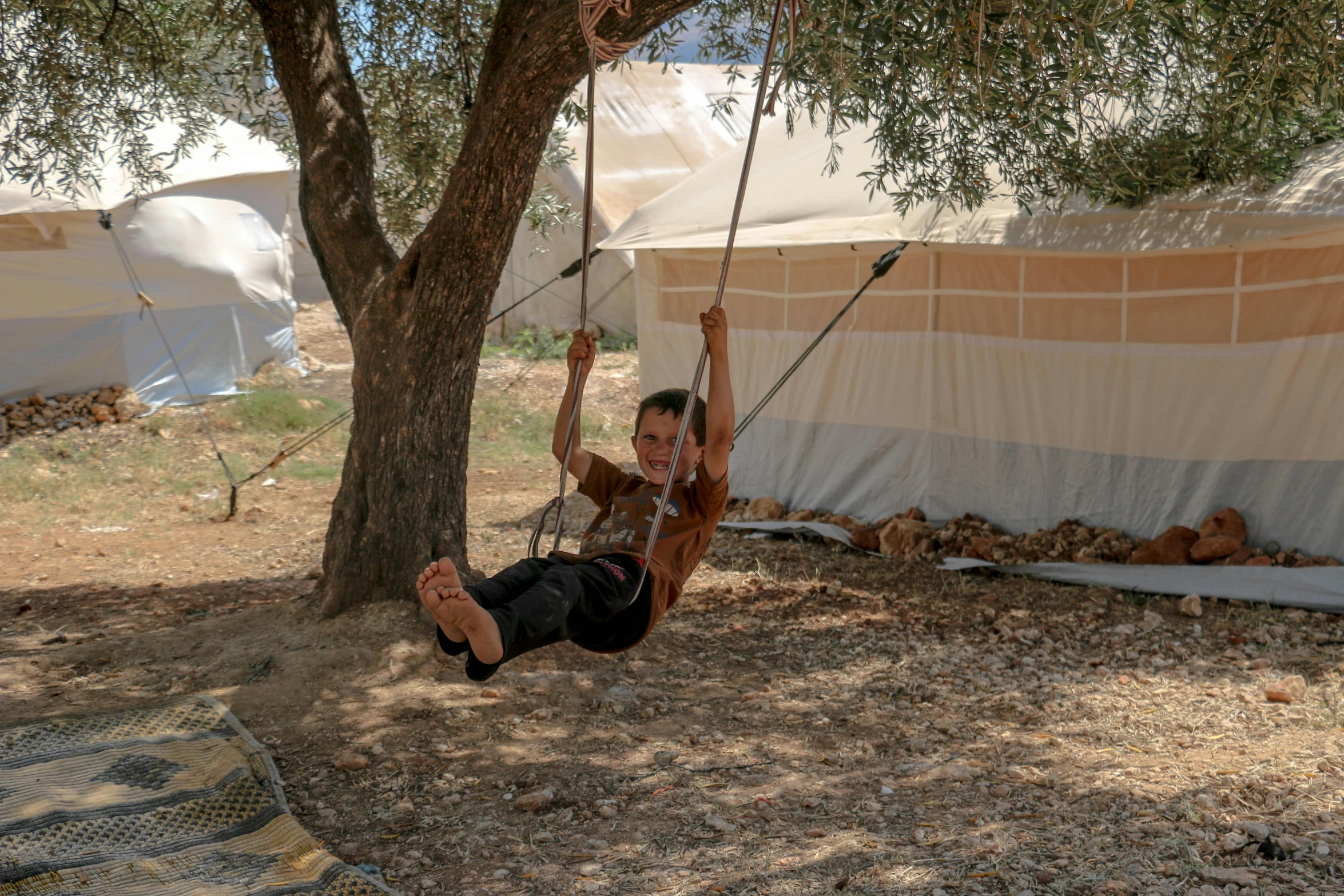 a man sitting on a swing next to a tree, hurufiyya, tents, refugees, kids playing, olive trees