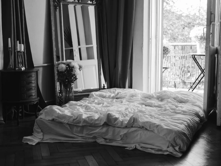 a bed sitting in a bedroom next to a window, a black and white photo, by Daniel Gelon, baroque, bed of flowers on floor, covered with blanket, with a mirror, uploaded