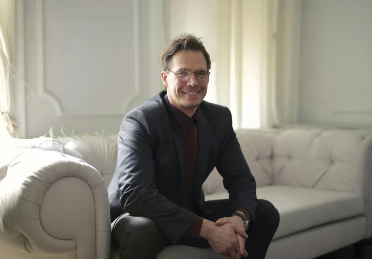 a man sitting on a couch in a living room, wearing a suit and glasses, julian ope, smiling man, in white room