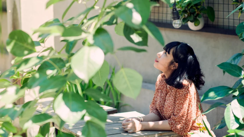 a woman sitting at a table in a garden, inspired by Yuko Tatsushima, unsplash, thoughtful pose, sunny day time, looking upward, greenery