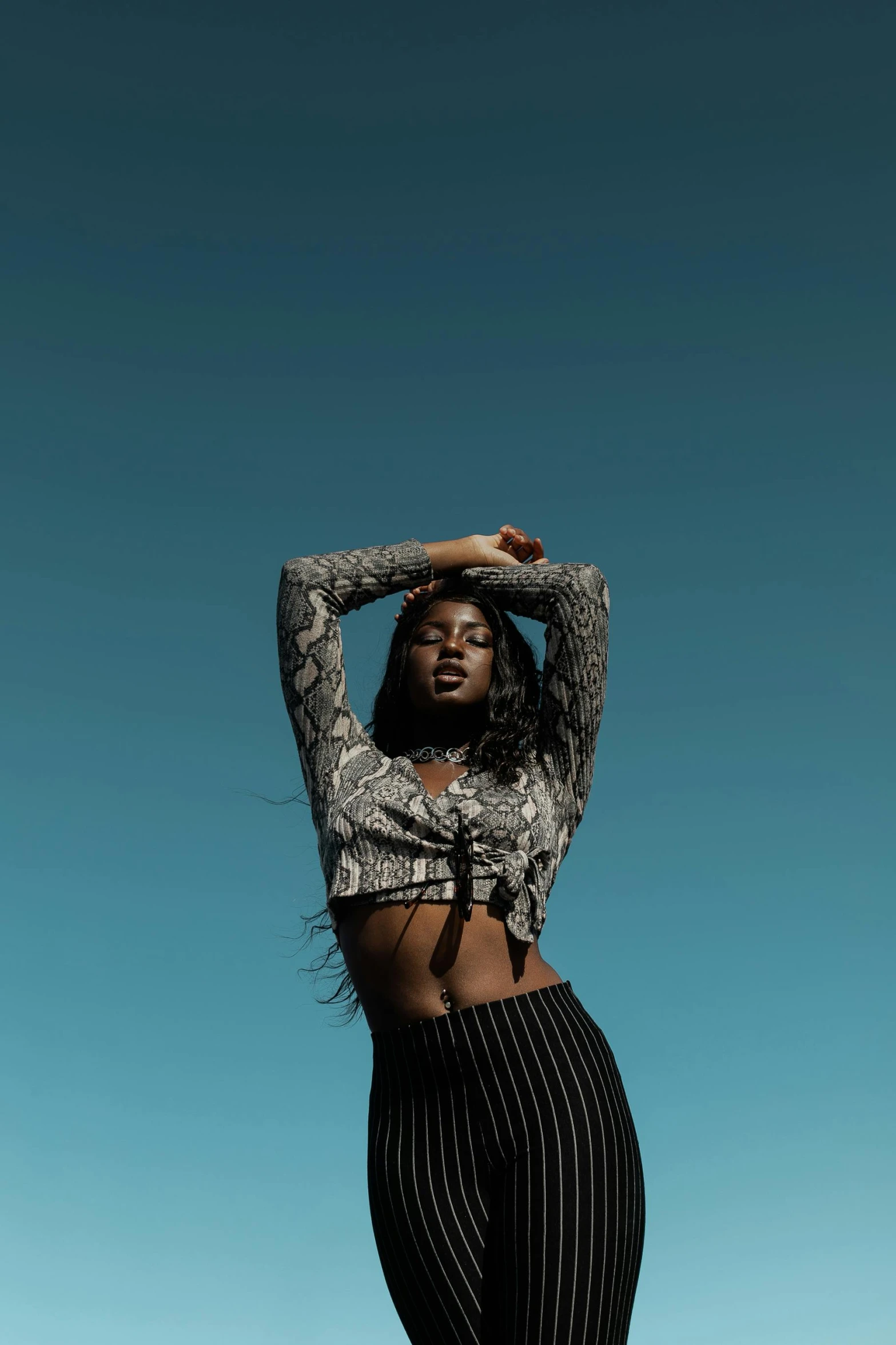 a woman standing in front of a blue sky, by Gavin Hamilton, trending on pexels, afrofuturism, wearing a sexy cropped top, patterned clothing, long windblown black hair, beauty expressive pose