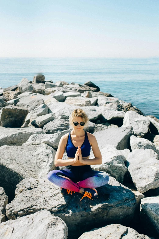 a woman sitting on top of a rock next to the ocean, wearing fitness gear, chakras, in barcelona, praying posture