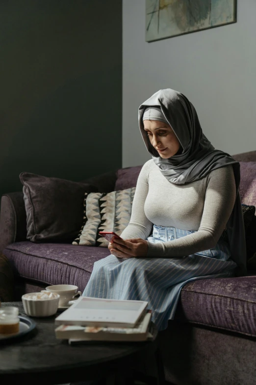 a woman sitting on a couch using a cell phone, inspired by Maryam Hashemi, hurufiyya, doing a prayer, looking serious, grey, head to waist