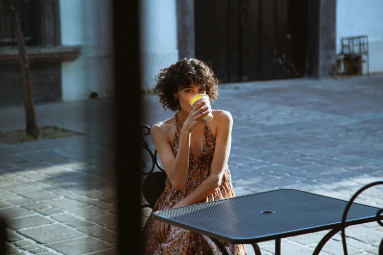 a woman sitting at a table eating an apple, by Daniel Seghers, pexels contest winner, wearing in a summer dress, woman drinking coffee, curly haired, wearing a yellow dress