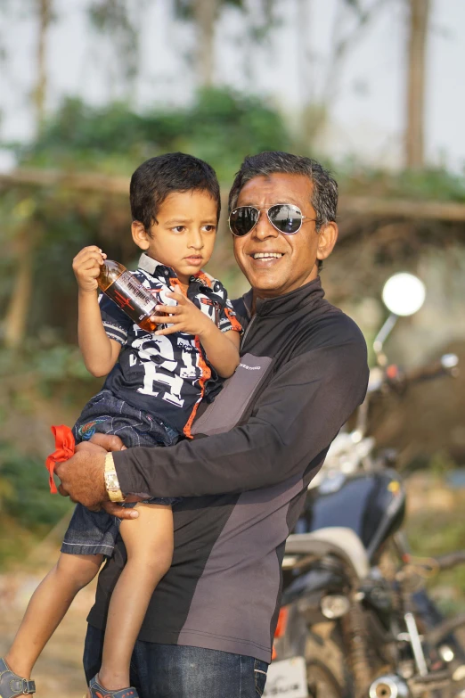 a man holding a child while standing next to a motorcycle, vinayak, wearing oakley sunglasses, portrait image, fatherly