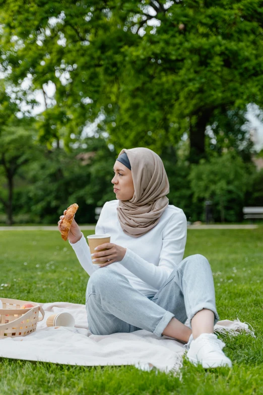 a woman sitting on a blanket eating a doughnut, by Adriaen Hanneman, shutterstock, white hijab, at a park, drink, eating garlic bread