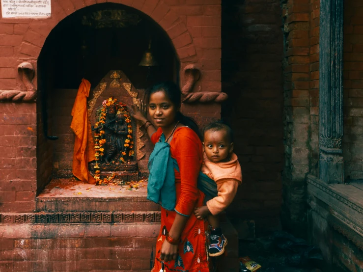 a woman holding a child in front of a statue, pexels contest winner, hurufiyya, nepali architecture buildings, wearing red and yellow clothes, in front of a fireplace, shrines