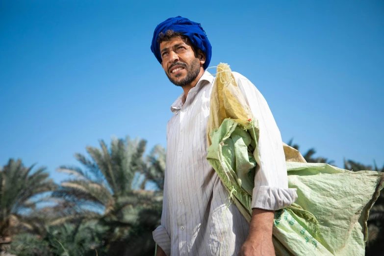 a man with a blue turban carrying a bag, by Scott M. Fischer, pexels contest winner, date palm trees, wearing farm clothes, portrait of bedouin d&d, a green