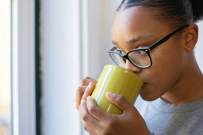 a woman drinking a cup of coffee in front of a window, trending on pexels, yellow and green scheme, nerdy black girl super hero, manuka, smelling good