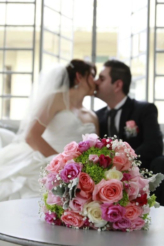 a bride and groom kissing in front of a wedding bouquet, sitting with flowers, indoor setting, pastel roses, full colour