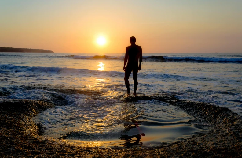 a man standing on top of a beach next to the ocean, by Jessie Algie, pexels contest winner, bathing in light, sun setting, standing in a shallow river, surfing