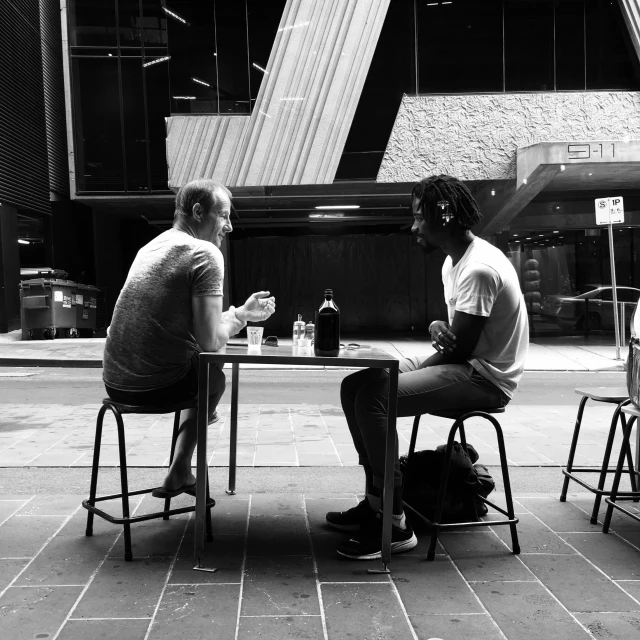 a black and white photo of two men sitting at a table, by Lee Loughridge, pexels contest winner, in chippendale sydney, in a square, taken on iphone 14 pro, talking