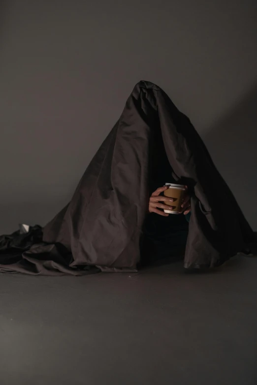 a person sitting under a blanket holding a cup, by Adam Chmielowski, conceptual art, pyramid head, black cloth, poop, promotional image