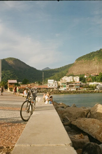 a man riding a bike down a sidewalk next to a body of water, a picture, inspired by Thomas Struth, trending on unsplash, rio de janeiro, village square, rocky beach, medium format. soft light