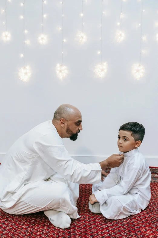 a man helping a young boy put on his shoes, by Sheikh Hamdullah, emitting light ornaments, wearing white pajamas, backdrop, professional photo