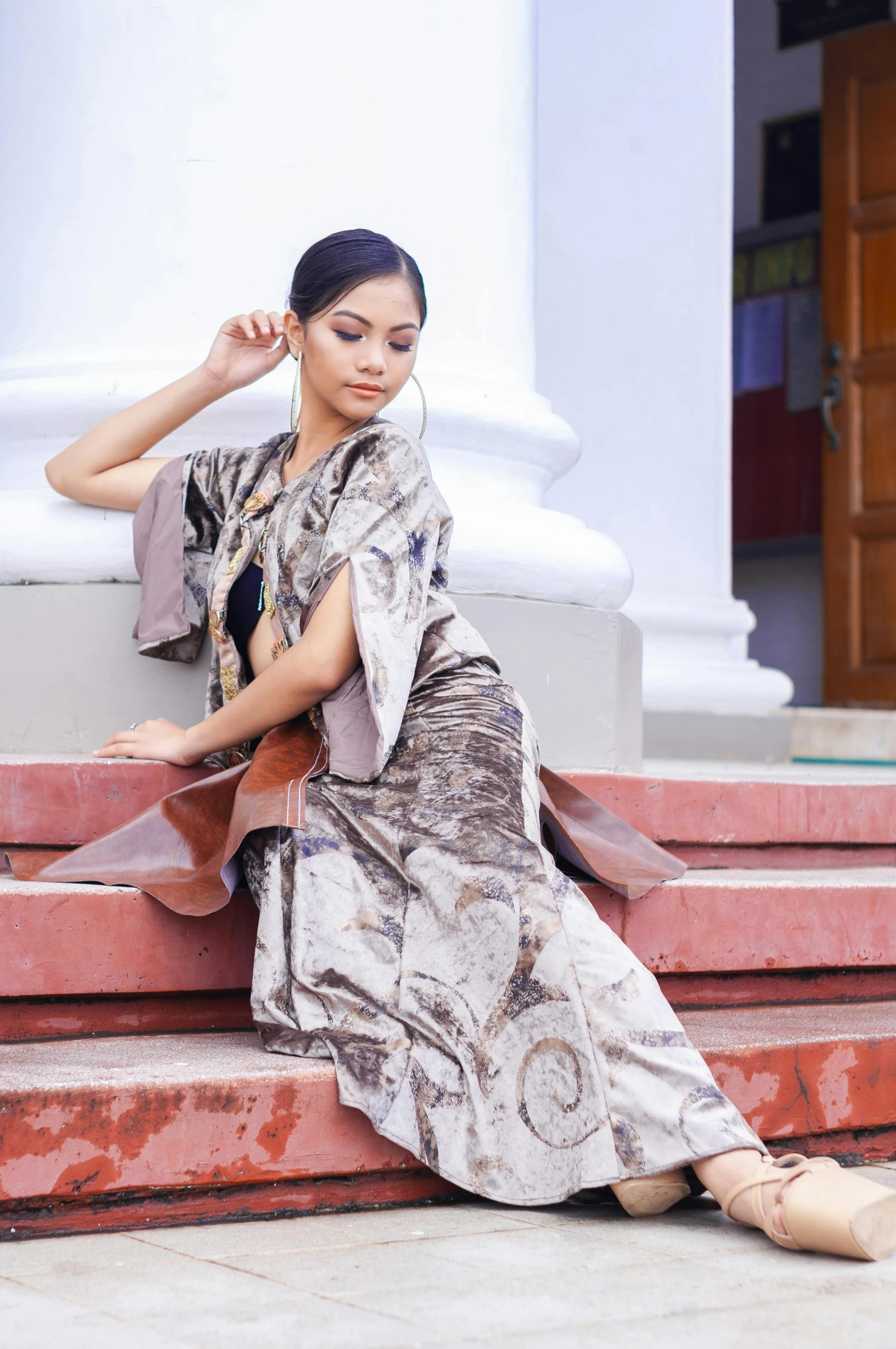 a woman sitting on the steps of a building, inspired by Sasha Putrya, arabesque, boho poncho and long maxi dress, batik, at a fashion shoot, classic beauty