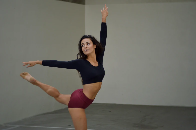 a woman standing on one leg in a dance pose, inspired by Elizabeth Polunin, pexels contest winner, arabesque, looking to the side off camera, cropped red yoga short, yael shelbia, physical : tinyest midriff ever