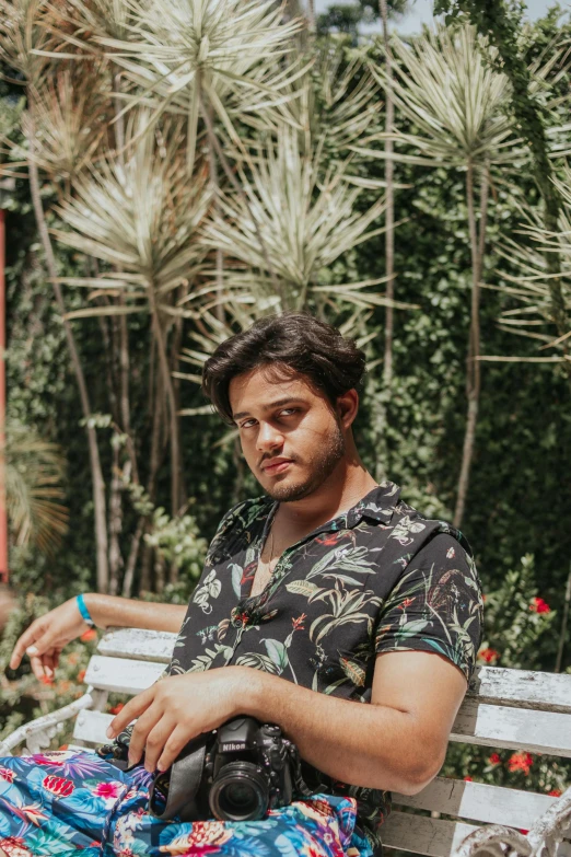 a man sitting on a bench with a camera, palm trees in the background, diego fernandez, profile image, patterned clothing