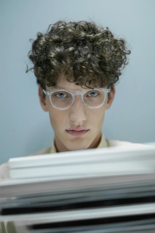 a young man sitting in front of a pile of papers, an album cover, inspired by Leo Leuppi, trending on pexels, small square glasses, curly haired, close-up portrait film still, looking into a mirror