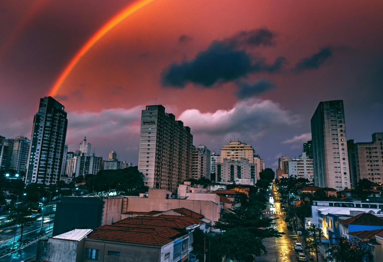 a rainbow shines in the sky over a city, an album cover, by Joze Ciuha, pexels contest winner, brazilian, ominous, slide show, red clouds