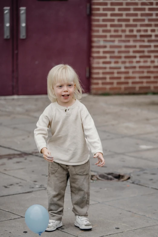 a little girl standing next to a blue balloon, wear's beige shirt, on sidewalk, paul barson, with hands in pockets