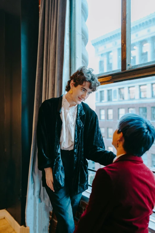 a man standing next to a woman near a window, by Nina Hamnett, trending on unsplash, finn wolfhard, wearing a luxurious velvet robe, in new york city, smiling at each other