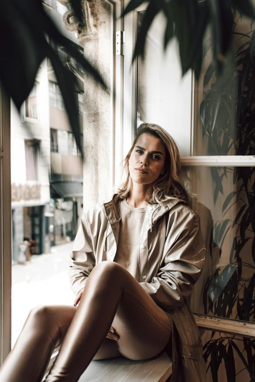 a woman sitting on a window sill next to a plant, a picture, inspired by Elsa Bleda, trending on unsplash, light brown trenchcoat, handsome girl, samara weaving, confident looking