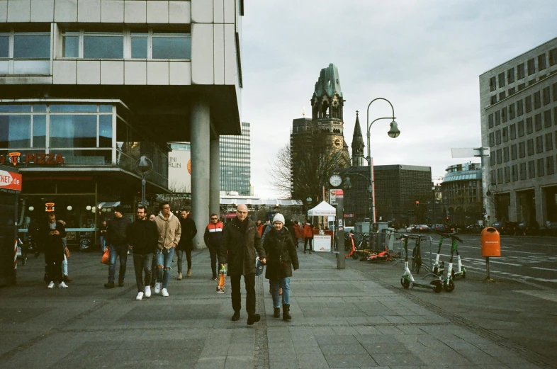 a group of people walking down a street next to tall buildings, an album cover, inspired by Thomas Struth, pexels contest winner, happening, palast der republik in berlin, church in the background, terminal, 16k resolution:0.6|people