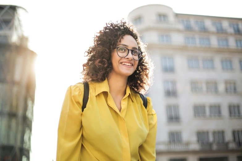 a woman with glasses standing in front of a building, wavy hair yellow theme, walking to work, smiling with confidence, schools