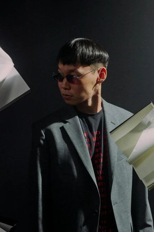 a man in a suit holding a piece of paper, an album cover, inspired by Fei Danxu, neo-dada, futuristic sunglasses, non binary model, asian man, standing in a dimly lit room