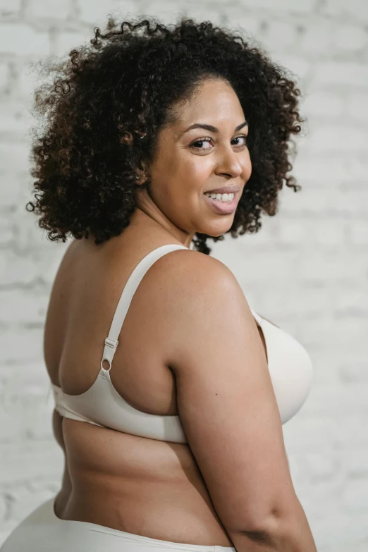 a woman in a white bra top standing in front of a brick wall, detailed product image, showing curves, person in foreground, d-cup