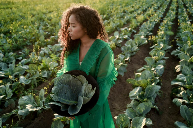 a woman standing in a field holding a hat, by Jessie Algie, pexels contest winner, renaissance, lettuce, nathalie emmanuel, green robes, confident holding vegetables
