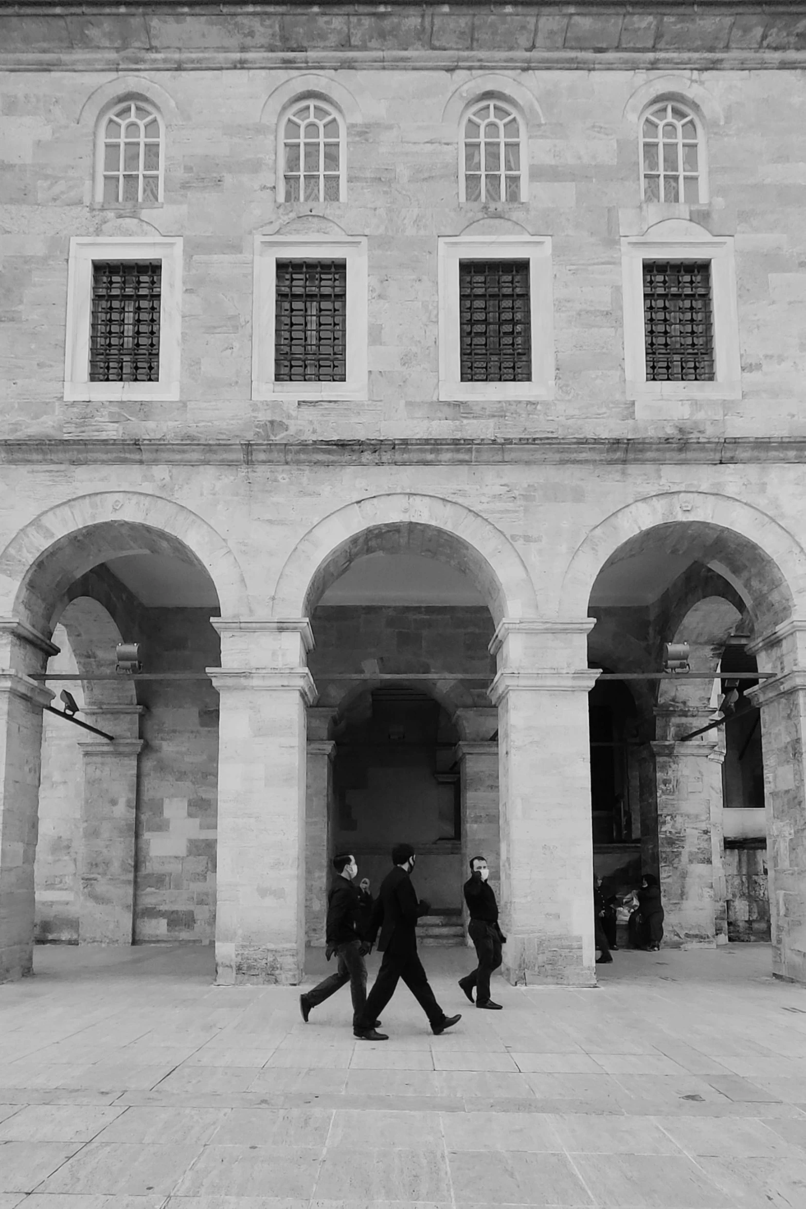 a black and white photo of two people walking in front of a building, a black and white photo, by Constantine Andreou, unsplash, romanesque, damascus, guards, people walking, threes