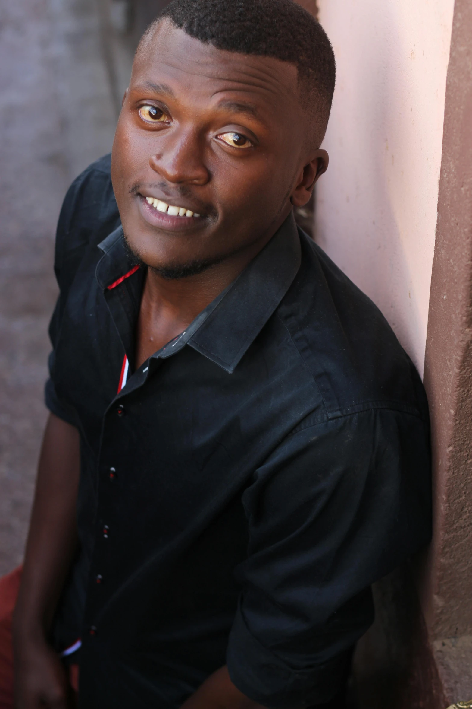 a man in a black shirt leaning against a wall, les nabis, dark skin tone, acting headshot, coloured, very kenyan