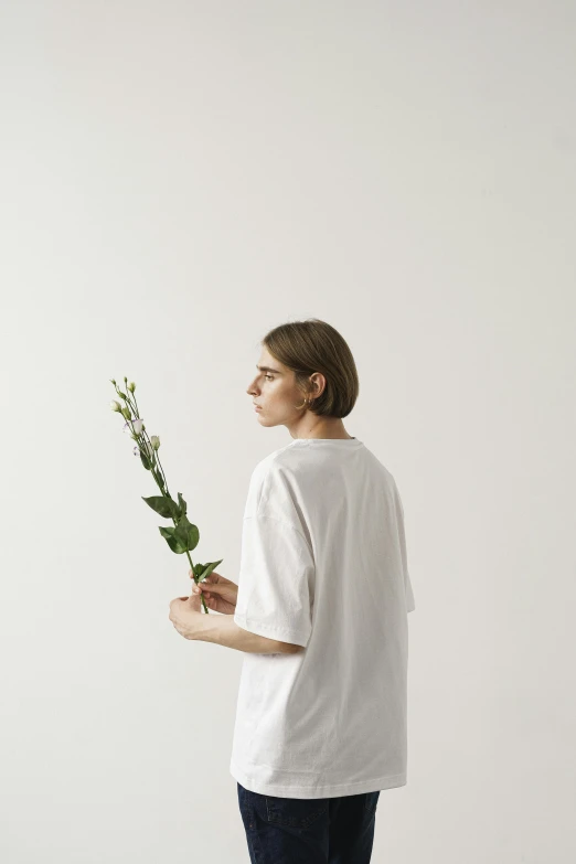 a man standing in front of a white wall holding a flower, by Anna Boch, minimalism, white tshirt, queer woman, ignant, flowers and stems